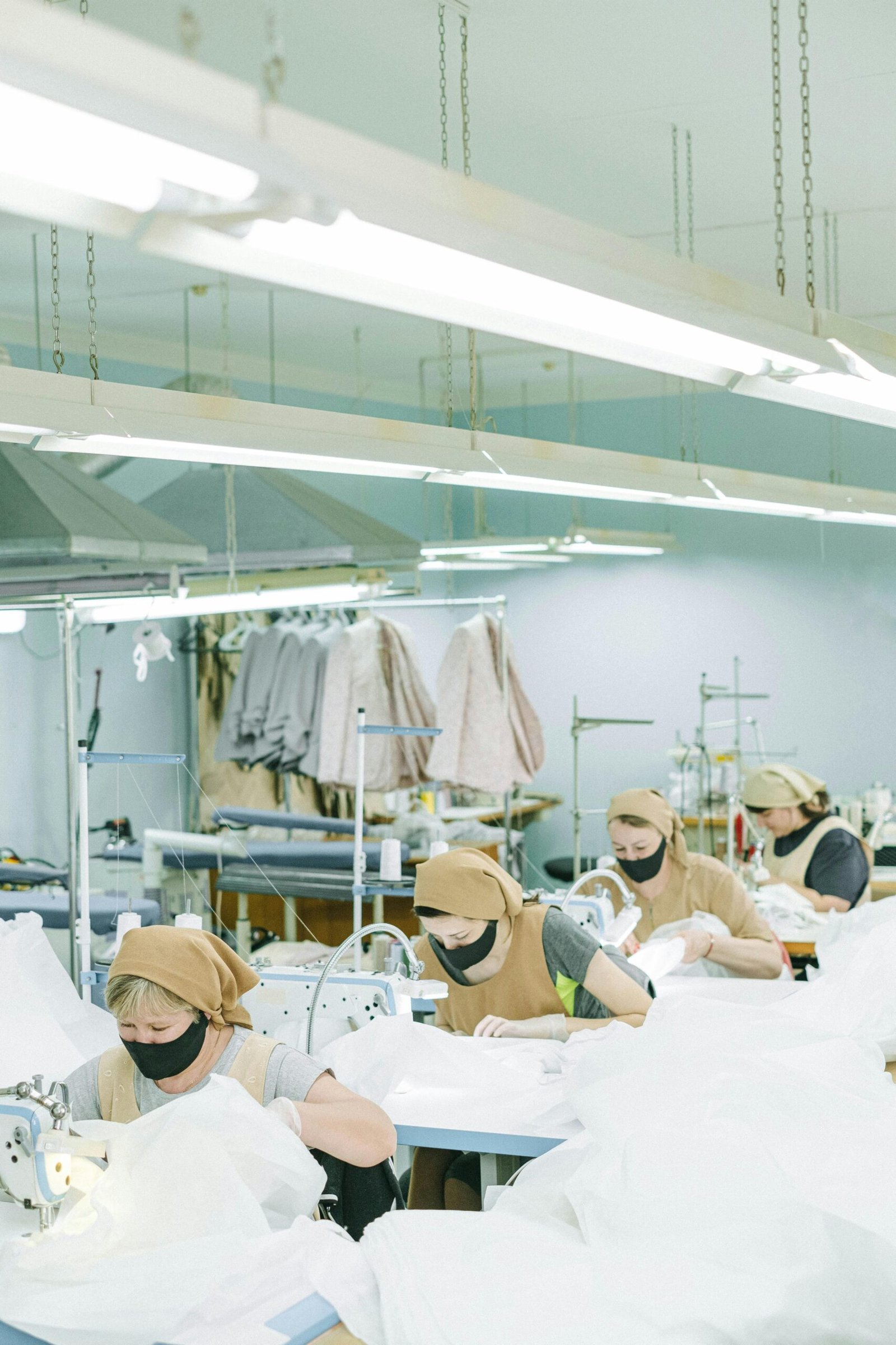 Women sewing with machines in a textile factory, wearing face masks and headscarves.